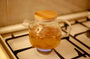 A clear glass teapot with a round bamboo lid sitting directly over a lit blue gas stove flame, containing warm amber herbal tea with visible loose dried botanicals steeping inside as steam condensation forms near the top rim.