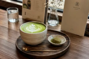 A high-angle shot captures a dark round wooden tray set on a warm wooden tabletop, presenting a large, speckled beige ceramic bowl filled with a matcha latte featuring a multi-layered tulip foam art design. Beside the bowl on the tray rests a small white dish holding a single green matcha mochi dusted with fine matcha powder, accompanied by a slim gold spoon placed in the foreground. In the soft-focused background, a clear glass vase with delicate dried baby's breath flowers, a glass of water, and paper menu cards—including one branded "Matchaya Kyoto"—are arranged against a bright window setup.