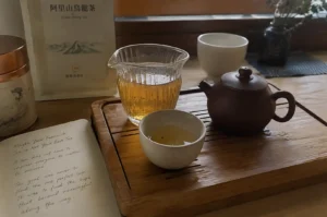 A cozy, eye-level tea preparation scene features a small dark brown clay Yixing teapot, a ribbed clear glass fairness pitcher filled with golden tea, and a small speckled ceramic teacup placed on a slotted wooden tea tray. To the left rests an open book displaying handwritten cursive text, alongside a copper-lidded tea canister and a paper pouch labeled "Alishan Oolong Tea" in Chinese characters, with a dark glass vase and a white ceramic cup softly positioned in the background.