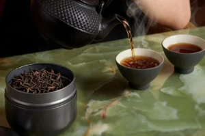 A dark, textured cast-iron teapot pours a hot stream of dark amber tea into one of two small, dark ceramic teacups set upon a polished green marble table. Wisps of steam rise from the cup as it fills, while a dark metallic tea canister filled with loose-leaf black tea sits open in the foreground.
