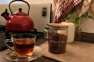 Set on a kitchen countertop, a clear glass cup on a saucer holds hot, amber-colored tea with a small brass spoon resting beside it. To the right on a linen mat sits a clear glass jar with a wooden lid, labeled "ROOIBOS TEA RED BUSH" and packed with loose reddish-brown tea leaves. A vibrant red kettle rests on the stovetop in the background alongside white storage canisters, a small glass vase with green plant cuttings, and red-and-white checkered dish towels hanging on the wall.