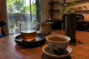 A close-up, eye-level shot captures a serene tea setup resting on a rich, dark wooden table inside a dimly lit café or teahouse. In the foreground on the right, a small porcelain teacup patterned with classic blue-and-white landscape motifs rests on a matching saucer, filled with warm, golden-brown tea. To the left sits a clear glass pitcher partially filled with amber tea, resting on a dark ceramic saucer, accompanied by a small, textured ceramic creamer or pitcher just behind it. On the far right, a tall, dark ceramic vase holds a small leafy green plant with vibrant leaves, catching subtle highlights. The background softly blurs into a warm indoor setting with wooden shelving and a large glass window that opens out onto lush green foliage illuminated by natural daylight.