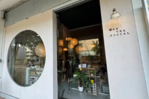 The storefront of Anteasocial features a clean, white minimalist facade with a large circular window on the left revealing warm interior shelves with teaware. On the right, black block lettering spells out "AN TEA SOCIAL" below a small, vintage-style sconce light. The glass entrance door opens into an inviting interior illuminated by warm, spherical paper lantern lights suspended at varying heights, with potted greenery framing the threshold.