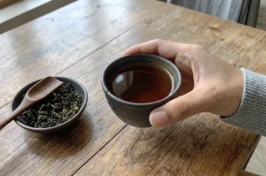 Person holding a dark ceramic tea cup filled with amber tea next to a small bowl of dried loose tea leaves on a rustic wooden table.