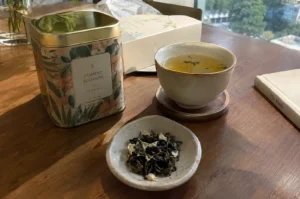 Jasmine Blossom tea tin, loose tea leaves, and a brewed cup of tea on a wooden table.