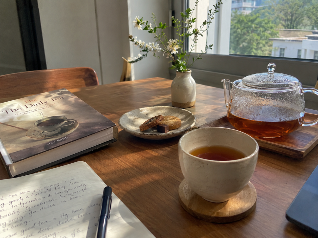 Sunlit wooden table set with tea, cookies, a book, notebook, and small flower vase.