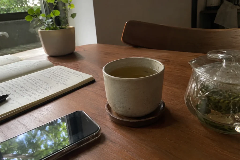 Ceramic tea cup, glass teapot, handwritten journal, and smartphone on a wooden study desk.