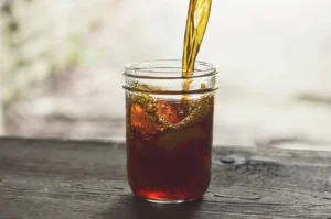 Iced tea beverage pouring into a mason jar with ice on a wooden table.
