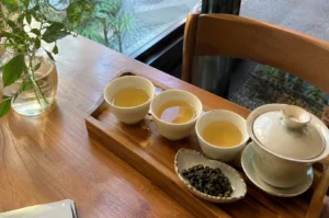 Traditional tea tasting set arranged on a wooden tray with three white cups of amber tea, a ceramic gaiwan, and rolled oolong tea leaves beside a small vase of greenery.