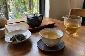 Traditional tea tasting set featuring loose tea leaves, a small dark clay teapot, a ceramic teacup filled with light amber tea, and a glass fairness pitcher on a wooden table.