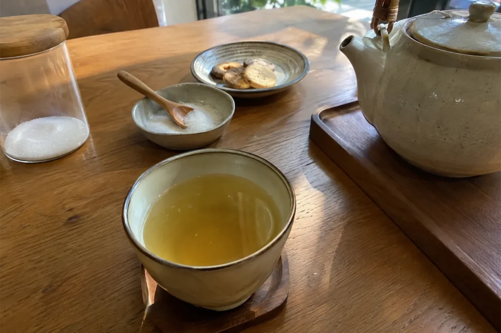 Wooden tabletop featuring a warm cup of herbal tea, a matching ceramic teapot on a tray, a small bowl of sugar with a wooden spoon, a glass jar, and a plate of biscuits.