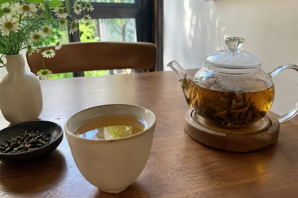A teapot of golden tea on a wooden coaster, a matching teacup, a bowl of loose leaves, and a vase of white flowers on a table.