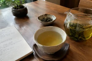 Traditional tea setup featuring a ceramic cup of brewed green tea, a glass teapot, loose tea leaves in a small dish, and a handwritten notebook on a wooden table.