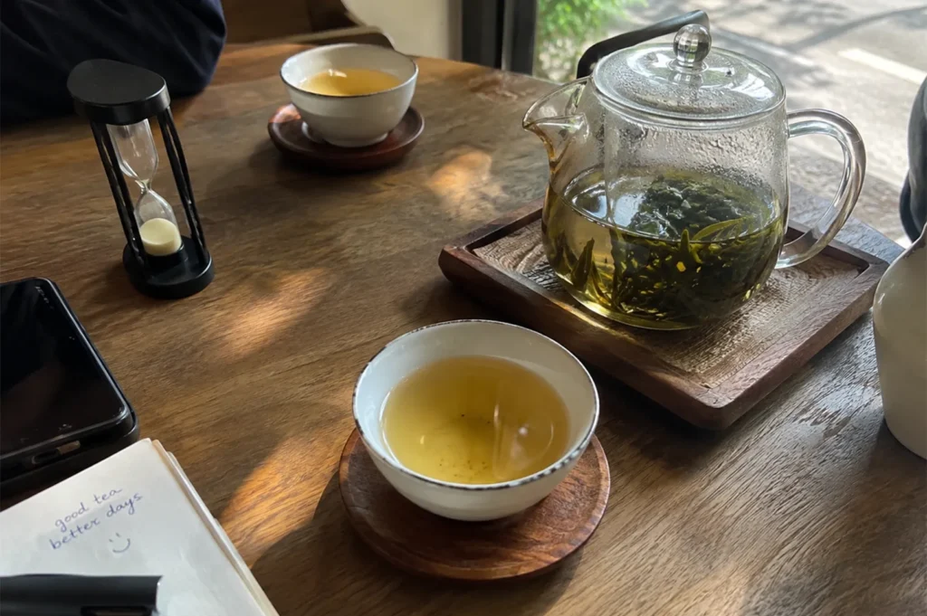 Clear glass teapot filled with steeping green tea paired with ceramic teacups and a sand timer on a wooden table.