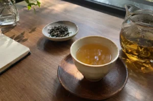 Minimalist tea ceremony arrangement with a ceramic cup of amber tea on a wooden saucer, a glass pitcher, dried tea leaves, and an open notebook on a wooden table.