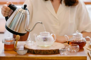 Person pouring hot water from a stainless steel gooseneck kettle into a clear glass teapot during a tea preparation ritual.
