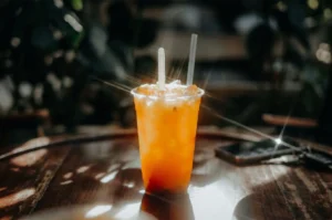 Clear plastic cup filled with an iced orange beverage and two straws resting on a dark wooden table with sun flares.