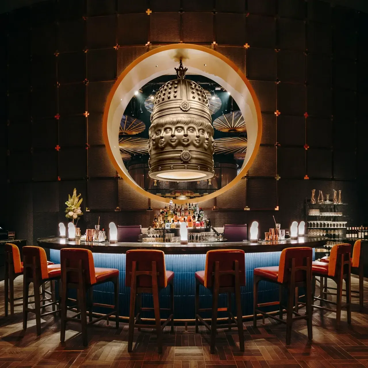 This upscale bar features a curved counter with orange barstools positioned beneath a large circular opening that reveals an ornate, multi-faced bronze bell. The dark, atmospheric space is accented by warm lighting and a textured wall, creating a sophisticated and mystical ambiance.