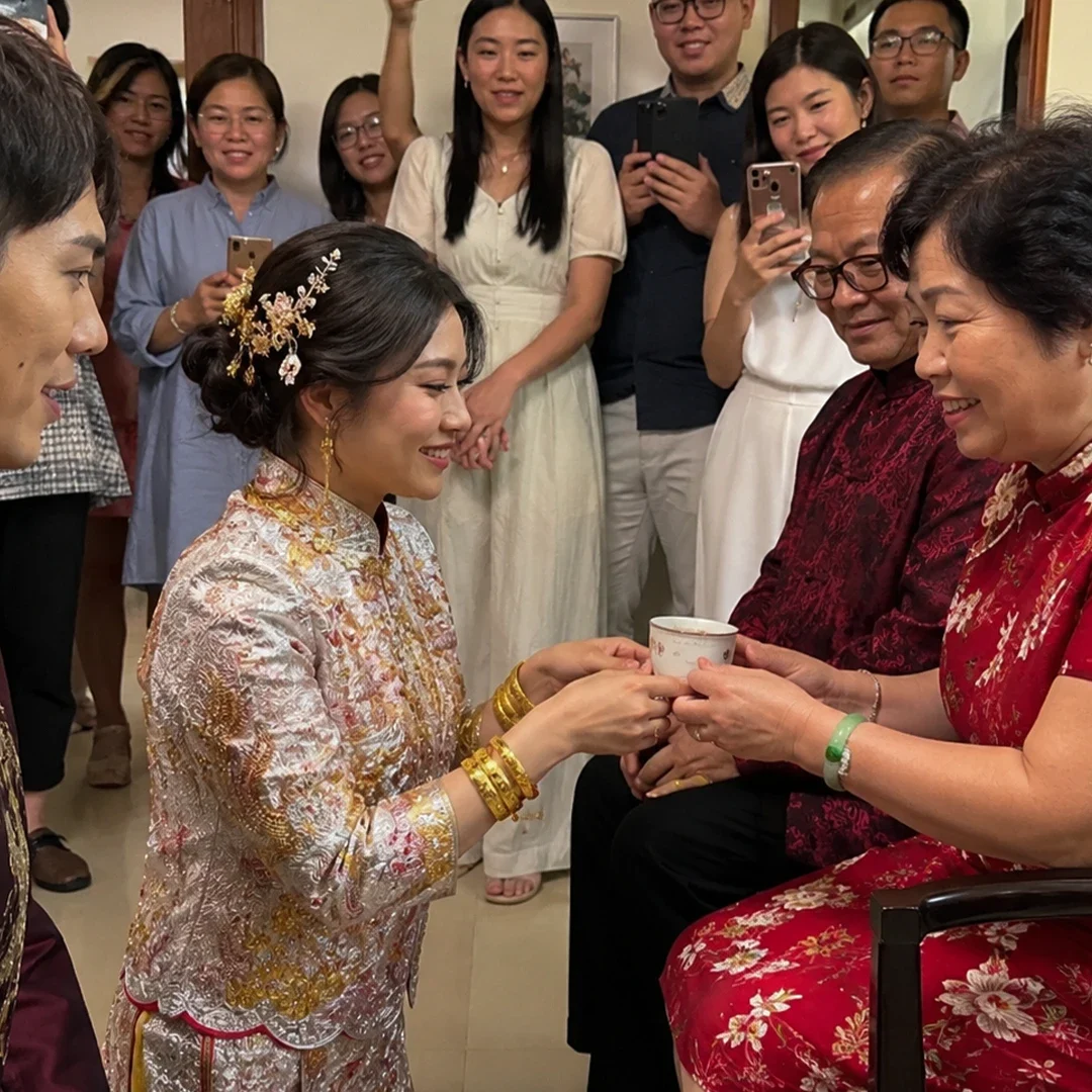 A bride in a white and gold embroidered Qun Kwa dress knees down, smiling as she presents a small white tea cup to an older woman wearing a red floral cheongsam dress. The bride wears heavy gold bangles on both wrists and an intricate gold floral hairpin in her updo, while the groom, dressed in a dark maroon embroidered tunic, stands close by her side. The older woman sits next to a man in glasses and a dark red shirt, accepting the tea with a smile. Behind them, a crowd of smiling family members stands in a doorway, holding up smartphones to capture the traditional ceremony beneath a red "Double Happiness" sign on the wall.