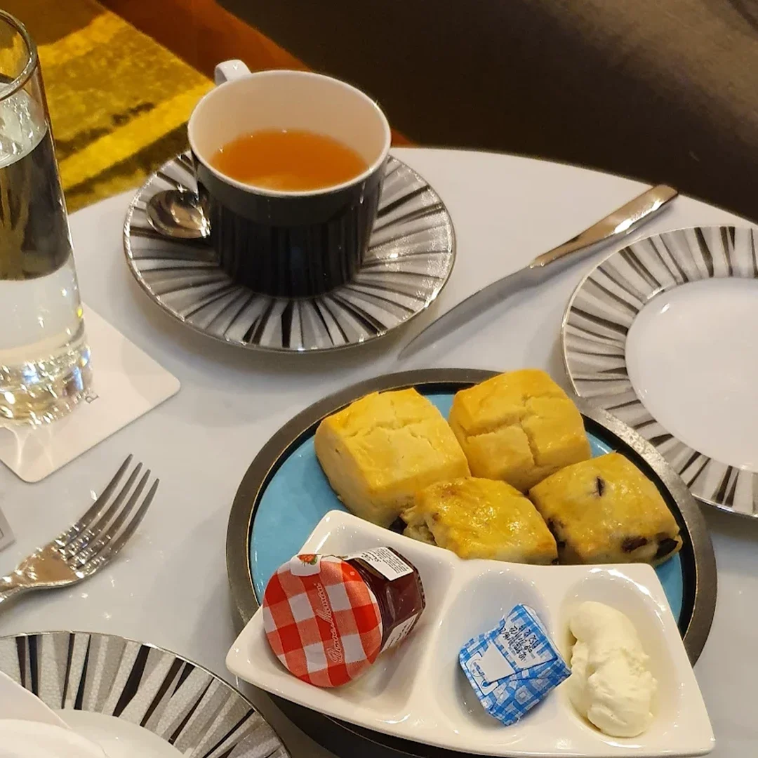 A close-up view of a high tea setting on a white table features a striped teacup and saucer paired with a plate of four golden-brown scones, served alongside a small dish containing a miniature jar of strawberry jam, a butter portion, and a dollop of clotted cream. A tall glass of water and a menu stand with a QR code are also visible on the table, which is set with a silver knife and fork, highlighting the refined details of the afternoon tea service.