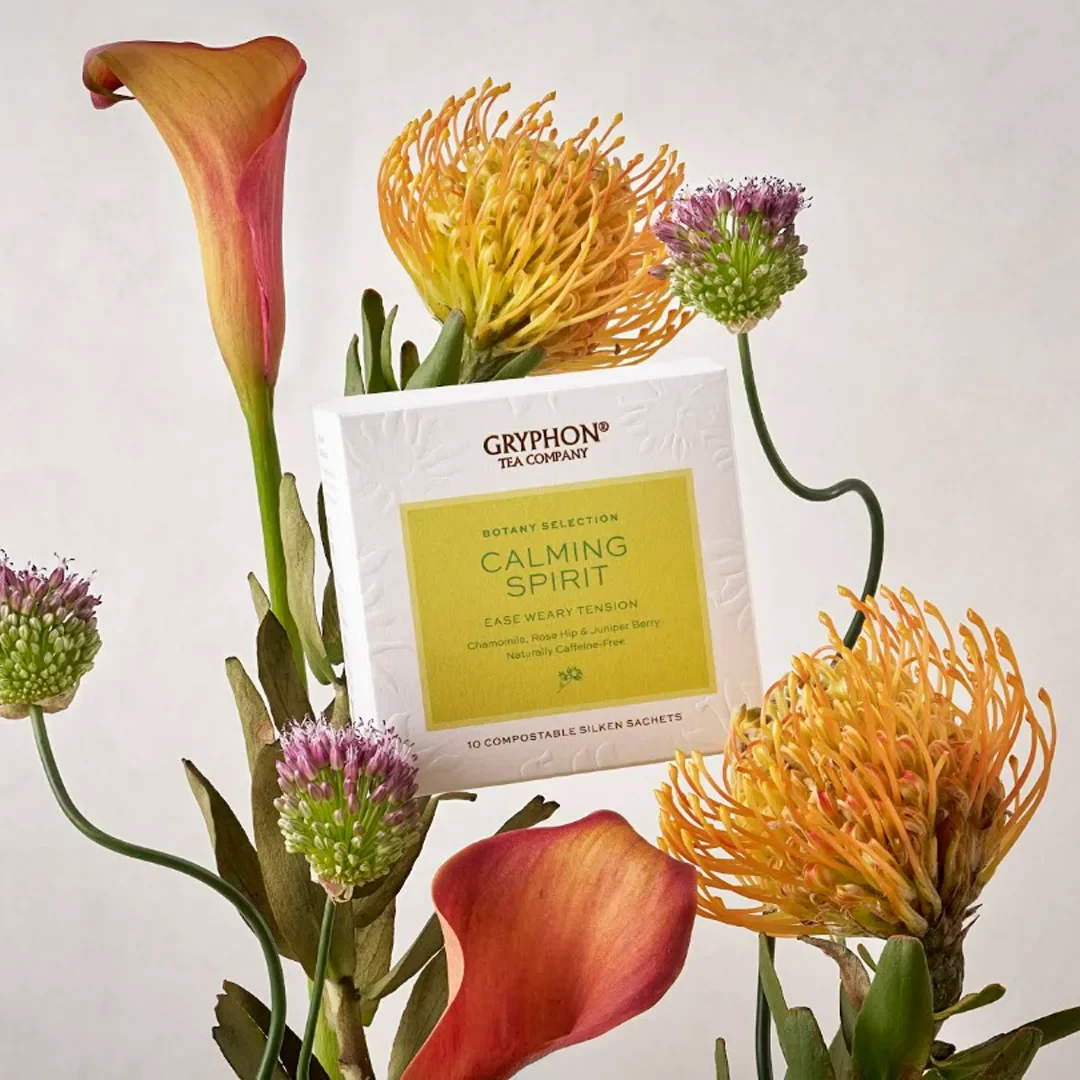 A close-up shot of the white box packaging for Gryphon Tea Company's "Calming Spirit" tea from their Botany Selection, nestled artistically amid a striking floral arrangement against a neutral beige background. The tea box features a lime-green central label detailing the blend as caffeine-free with chamomile, rose hip, and juniper berry. It is framed by a dynamic cluster of unique botanical stems, including two vibrant, orange-tipped yellow pincushion proteas, a pair of tall, elegant calla lilies in warm sunset shades of orange and pink, and several slender green stalks sporting round, spiky purple and green allium flower buds.