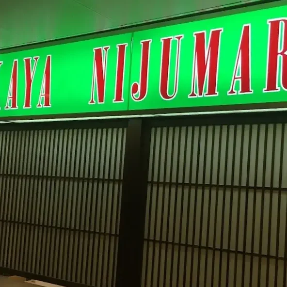 This image shows a brightly lit, neon green sign for a Japanese restaurant called "Izakaya Nijumaru," featuring bold red lettering with white outlines. Below the sign, traditional-style dark wooden lattice screens with frosted glass panels form a storefront or partition.