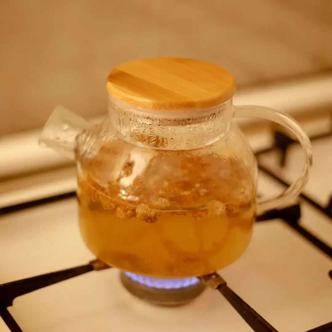 A clear glass teapot with a round bamboo lid sitting directly over a lit blue gas stove flame, containing warm amber herbal tea with visible loose dried botanicals steeping inside as steam condensation forms near the top rim.