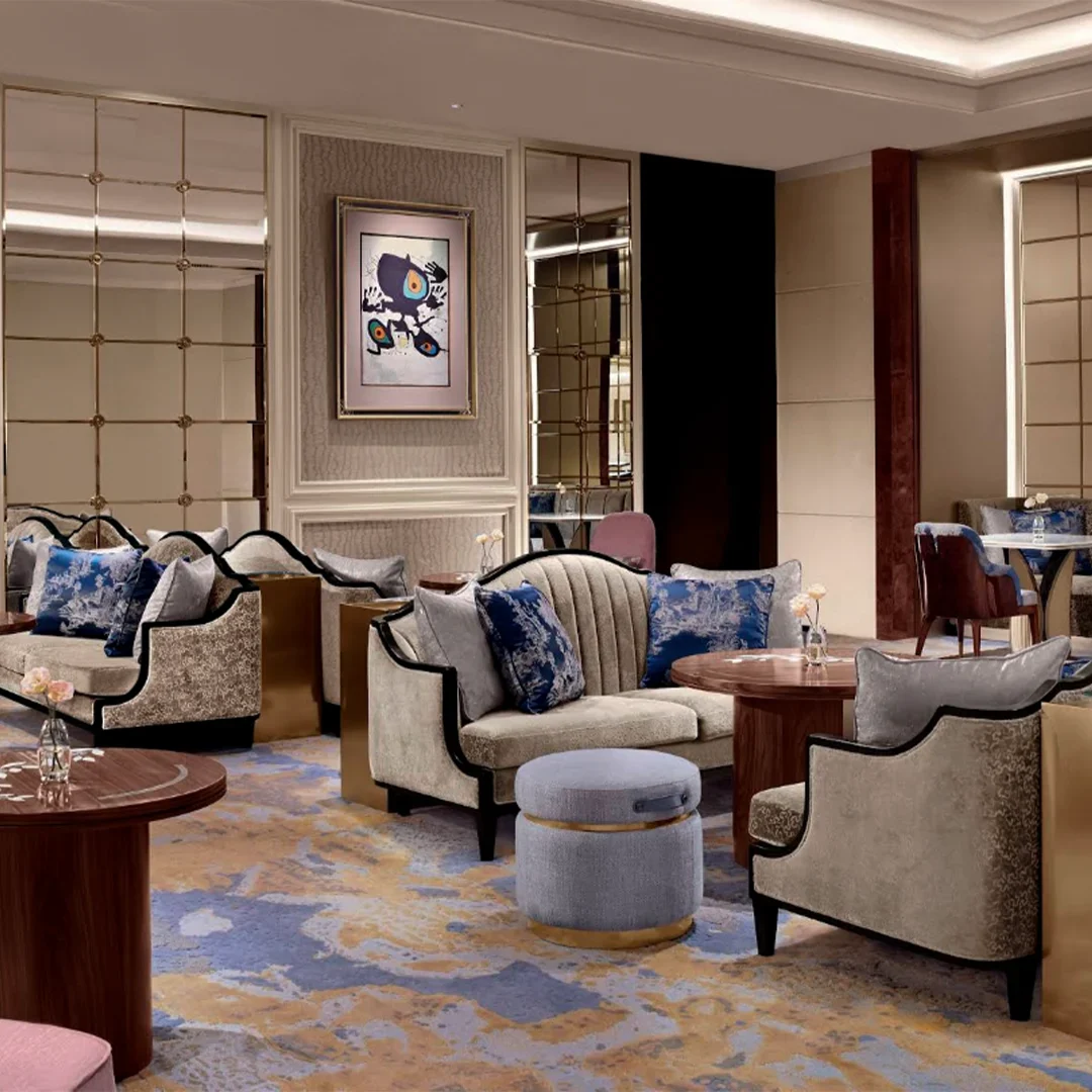 This wide shot captures an elegant and luxurious hotel lounge decorated in sophisticated muted tones, featuring plush seating arrangements spaced across a large, patterned carpet with swirling shades of blue, gold, and cream. The main seating area includes curved beige sofas adorned with deep blue and silver-patterned throw pillows, paired with small round wooden coffee tables, a low grey velvet ottoman with a gold base, and armchairs with soft pink velvet upholstery. The walls are finished in a neutral textured wallpaper, punctuated by large grid-patterned mirrors that reflect the room and modern abstract framed artwork, creating an inviting yet upscale ambiance.