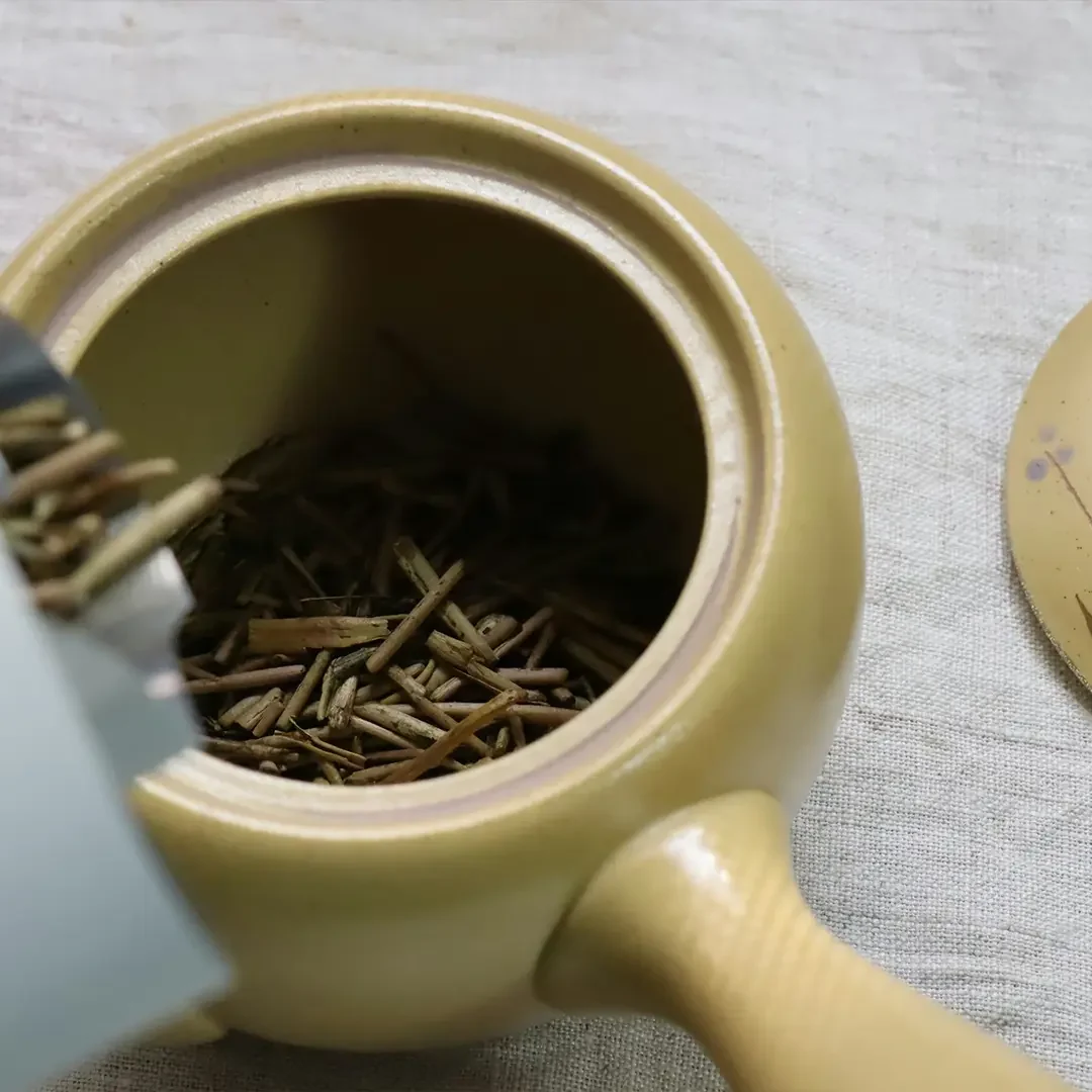 Dry, needle-like tea leaves are being poured from a light-colored paper bag into a yellow ceramic teapot, with the lid of the teapot resting nearby on a textured beige fabric surface.