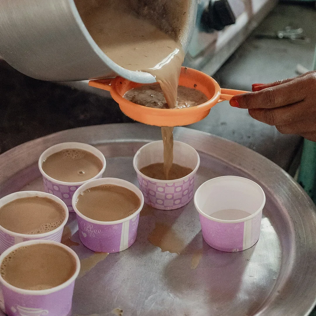 A person’s hand is seen straining hot, milky masala chai through a small orange plastic sieve from a large metal pot into several paper cups arranged on a metallic tray, illustrating the preparation process in a busy kitchen setting.