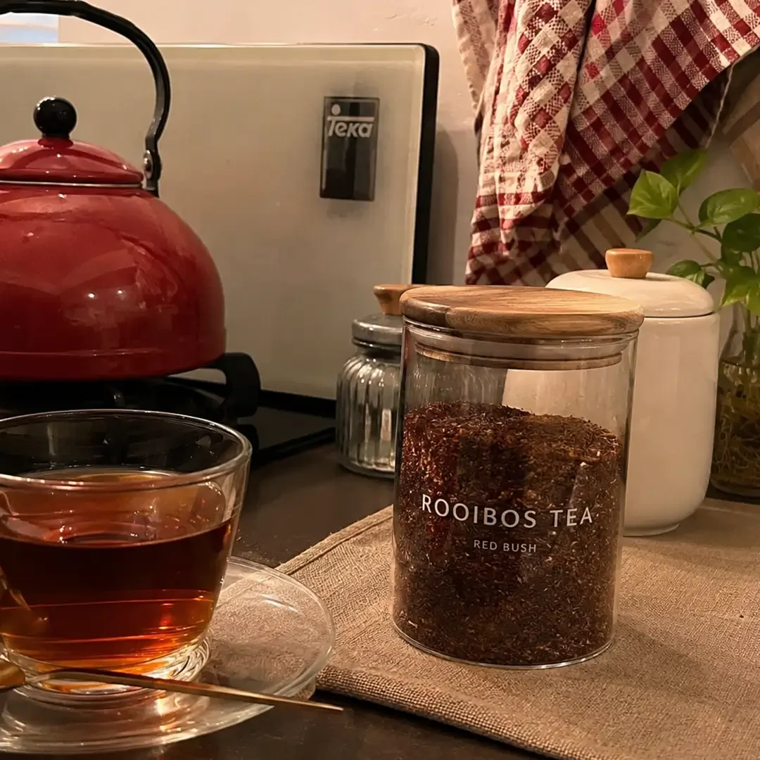 Set on a kitchen countertop, a clear glass cup on a saucer holds hot, amber-colored tea with a small brass spoon resting beside it. To the right on a linen mat sits a clear glass jar with a wooden lid, labeled "ROOIBOS TEA RED BUSH" and packed with loose reddish-brown tea leaves. A vibrant red kettle rests on the stovetop in the background alongside white storage canisters, a small glass vase with green plant cuttings, and red-and-white checkered dish towels hanging on the wall.