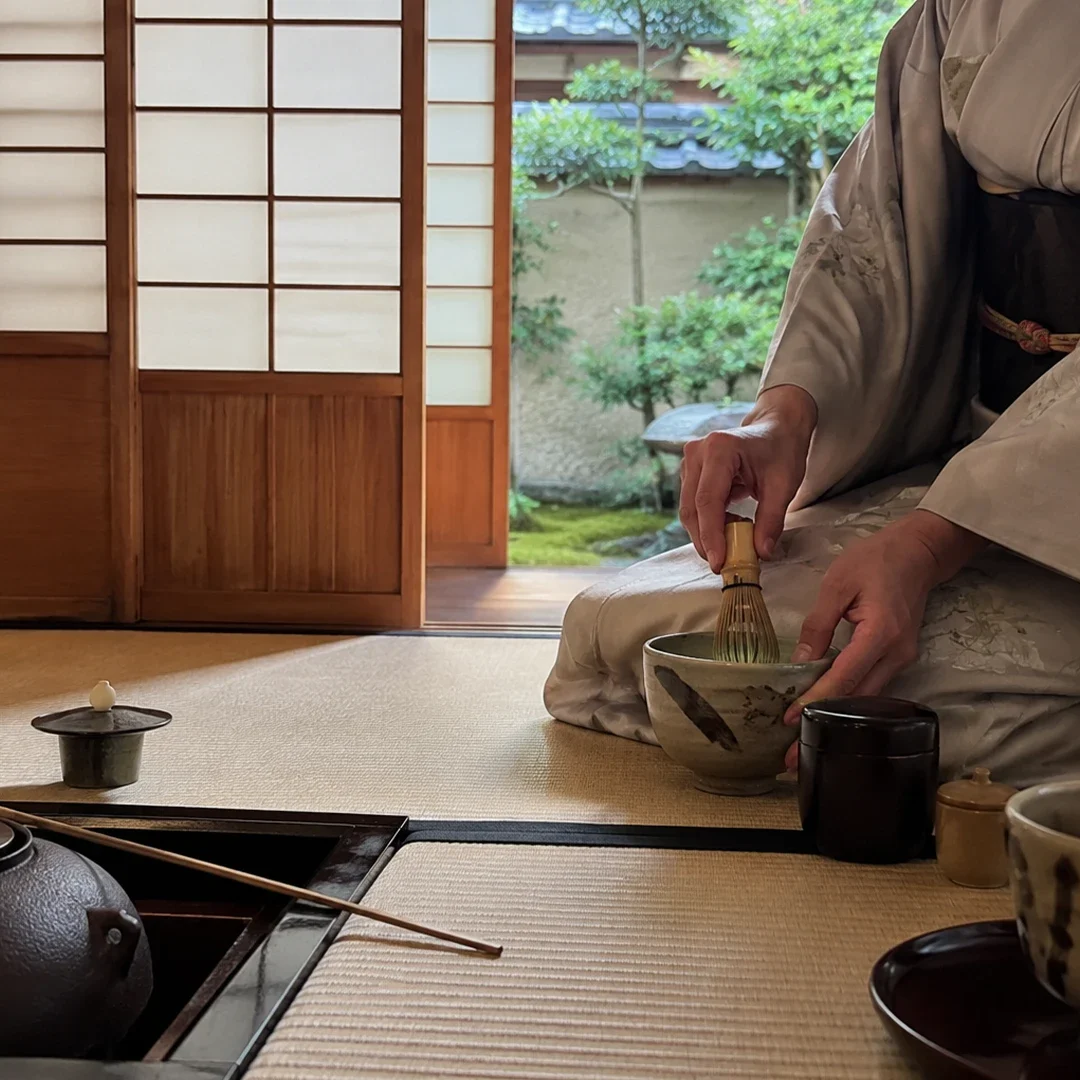A woman dressed in a soft-toned kimono kneels on a tatami mat in a traditional Japanese tea room, carefully whisking matcha in a rustic ceramic bowl with a bamboo chasen. Before her on the mat are various tools of the tea ceremony, including an iron kettle, a bamboo ladle, and a small tea caddy, all arranged with precision. In the background, open shoji sliding doors frame a serene view of a lush green garden, emphasizing the peaceful and focused atmosphere of the ceremony captured.