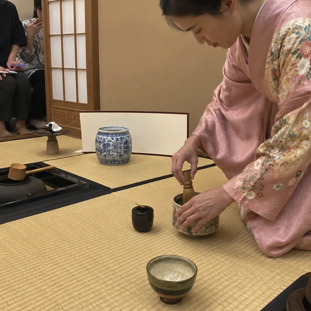 A Japanese tea ceremony unfolds on traditional tatami mat flooring, where a woman dressed in a delicate pink floral kimono kneels gracefully as she whisks matcha inside a large ceramic tea bowl. Various authentic tea utensils are meticulously arranged around her on the mats, including a blue and white patterned ceramic water jar, a black lacquer tea caddy, a wooden water ladle resting over a sunken hearth, and a small dark ceramic bowl in the foreground. In the background, partially separated by a paper shoji screen, a few guests sit on a low bench, quietly observing the practitioner's precise movements.