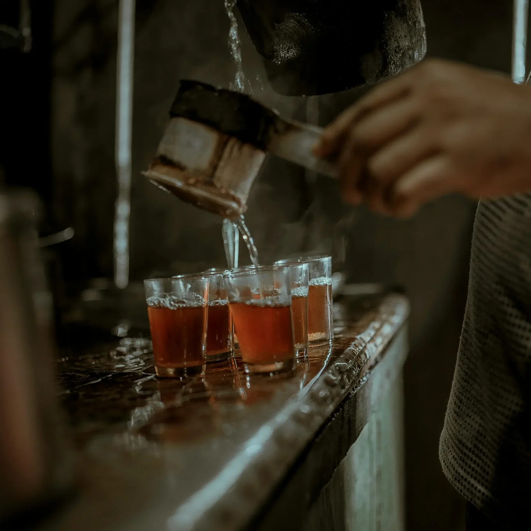 A person pours a dark, steaming liquid through a traditional cloth strainer into several small glasses lined up on a rustic counter. The scene is captured in a moody, low-light setting with a shallow depth of field that highlights the motion of the pour.