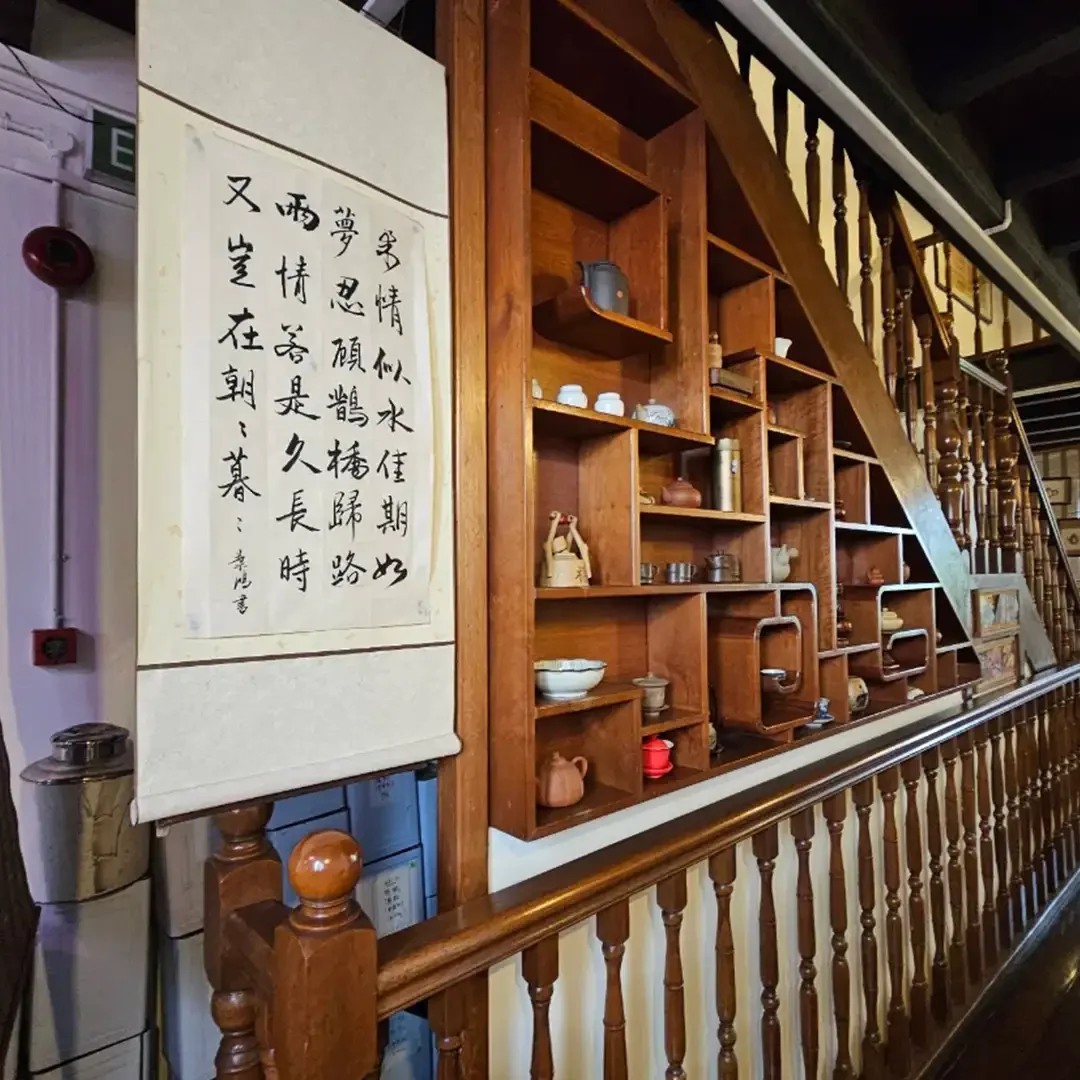 A wooden staircase with intricate balusters ascends along a wall in a traditional tea house setting. Mounting the wall is a vertical hanging paper scroll filled with handwritten Chinese calligraphy, positioned beside a custom-built, multi-tiered wooden display shelf holding various ceramic teapots and teaware.