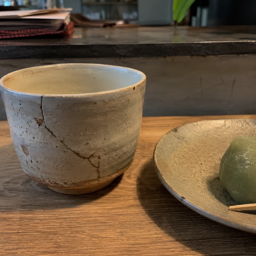 A close-up, eye-level shot features a striking example of kintsugi art on a wooden counter. On the left stands a light beige ceramic cup with a rustic, unglazed base, displaying prominent cracks beautifully mended with dark gold lacquer. To the right, a shallow, textured grey ceramic plate holds a single, round green mochi sweet with a small wooden pick resting against it. The background is dimly lit and out of focus, showing a dark stone counter lip and glimpses of papers and green leaves, keeping the emphasis on the repaired pottery.