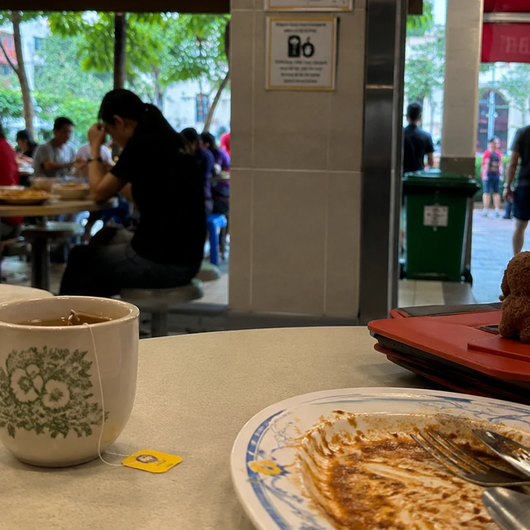 A close-up, high-angle shot of a minimalist tea setup on a light wooden surface. A clear glass teapot with a bamboo sleeve sits next to a small blue lidded ceramic cup (gaiwan). To the right, a matching blue cup featuring a whimsical line-art drawing of a face and the word "Magpie" rests on a black, hexagonal coaster. Natural sunlight casts sharp, dramatic shadows across the scene.