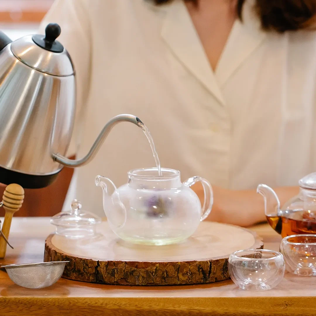Person pouring hot water from a stainless steel gooseneck kettle into a clear glass teapot during a tea preparation ritual.