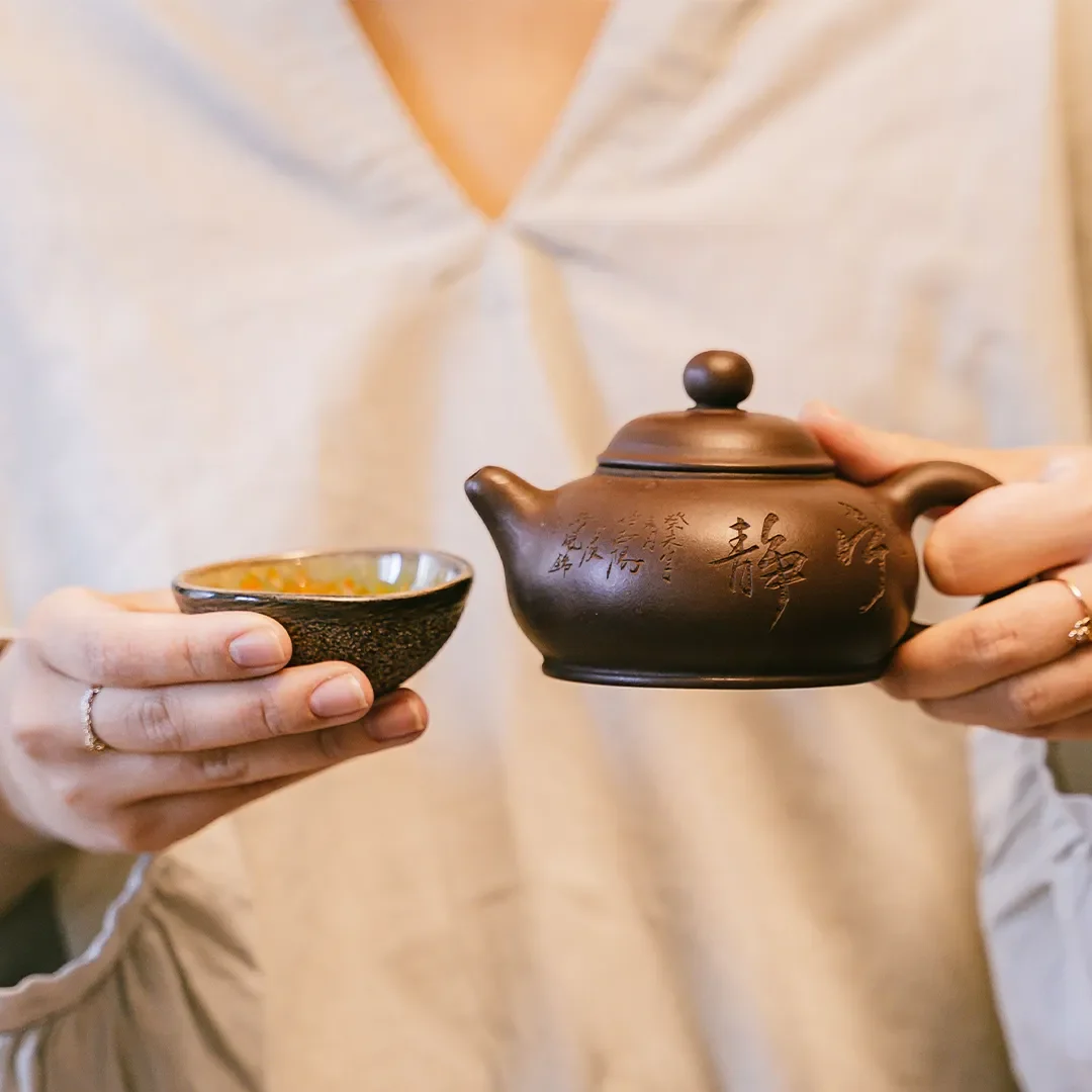 Person holding a small clay teapot with calligraphy and a ceramic teacup.