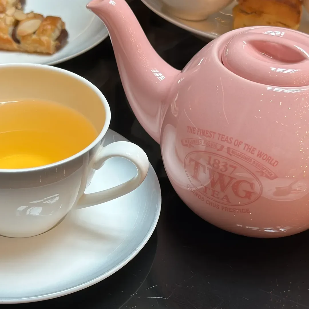 A close-up shot of a classic light pink TWG ceramic teapot and a filled teacup served with fresh pastries during an afternoon high tea.