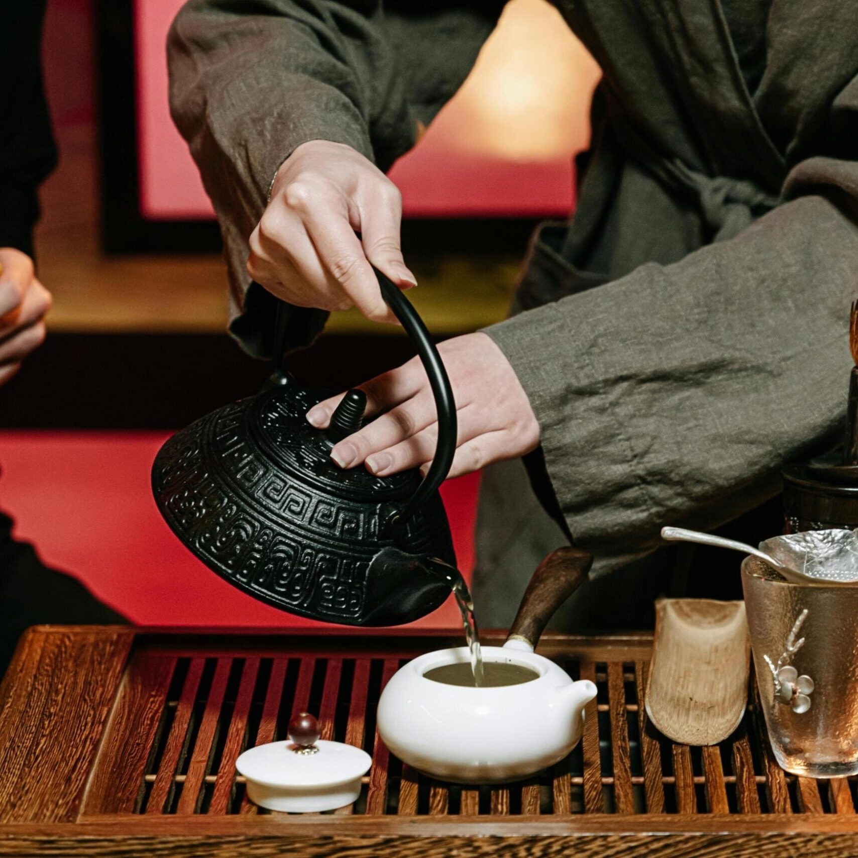 A person wearing a grey robe carefully pours hot water from a black, textured cast-iron kettle into a small white teapot during a traditional tea ceremony. The ritual takes place on a slotted wooden tray adorned with various tea accessories, including a glass pitcher, strainer, and bamboo tools, while another participant sits nearby.
