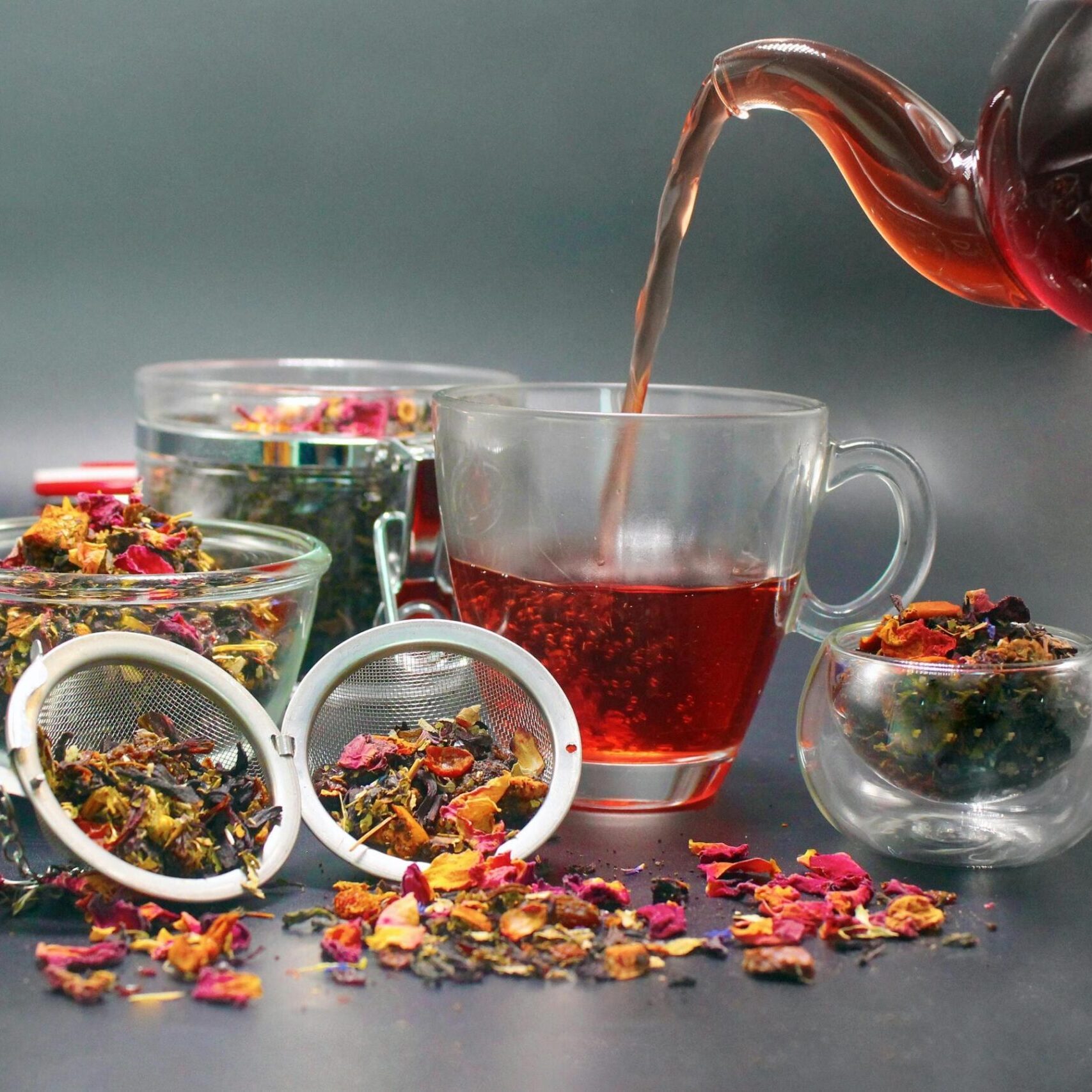 dark amber tea is poured from a glass teapot into a matching clear mug, surrounded by several glass bowls and mesh strainers filled with colorful loose-leaf tea. The vibrant blend of dried petals and herbs is scattered across the dark surface, highlighting the rich floral and botanical ingredients.