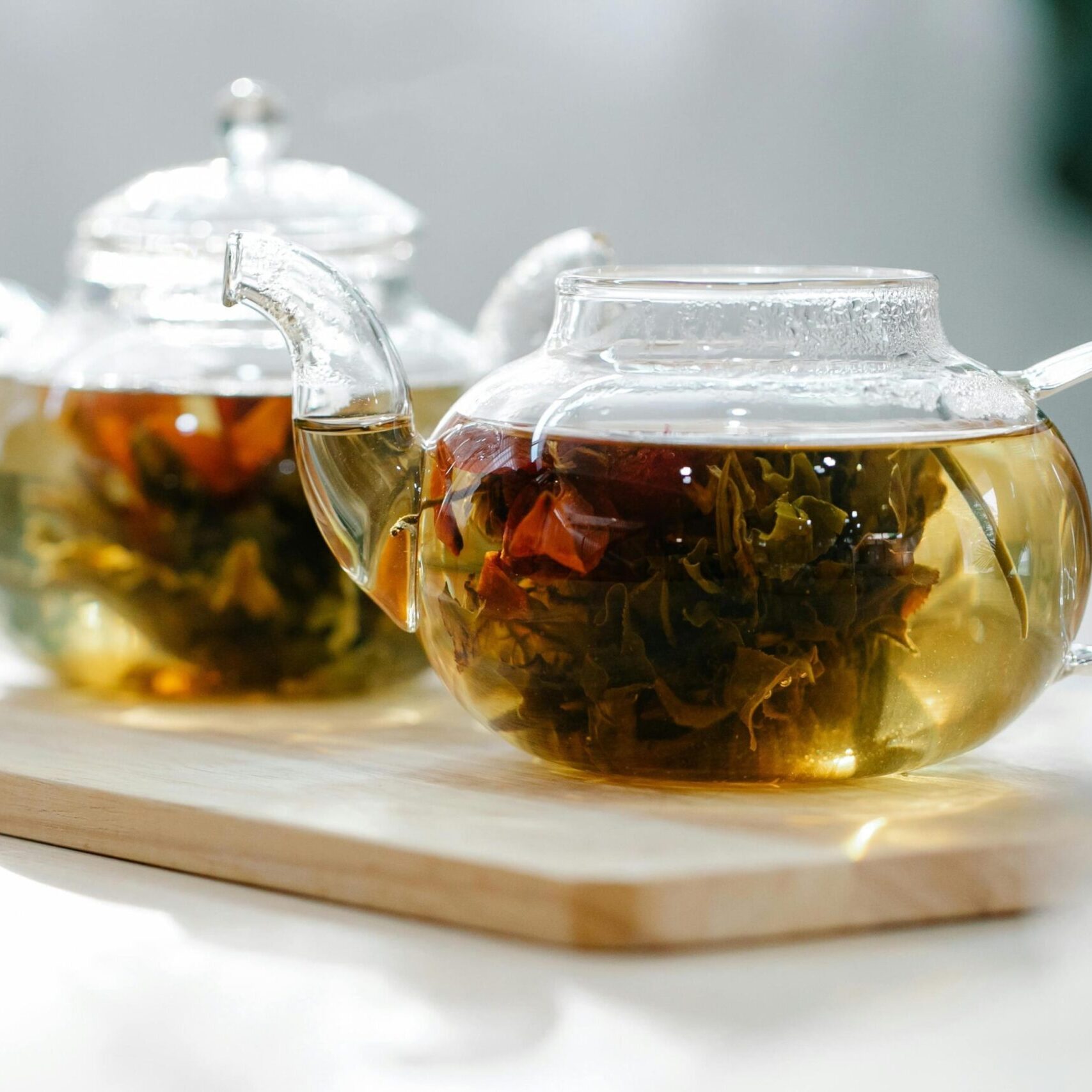 Two clear glass teapots rest on a light wooden tray, showcasing blooming tea flowers unfurling within the golden liquid. Bright natural light streams through the glass, illuminating the amber tea and creating a warm, serene atmosphere.