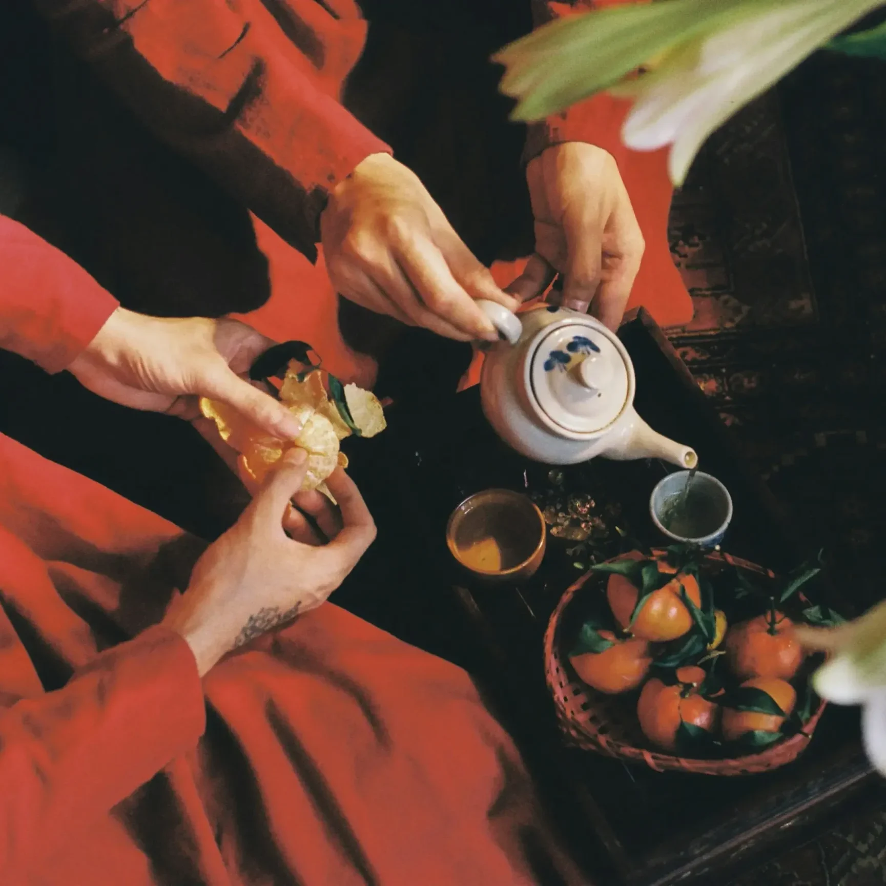 Two people wearing vibrant red garments share a traditional moment of tea and fruit over a patterned rug. One person carefully pours tea from a ceramic pot while the other peels a fresh tangerine.