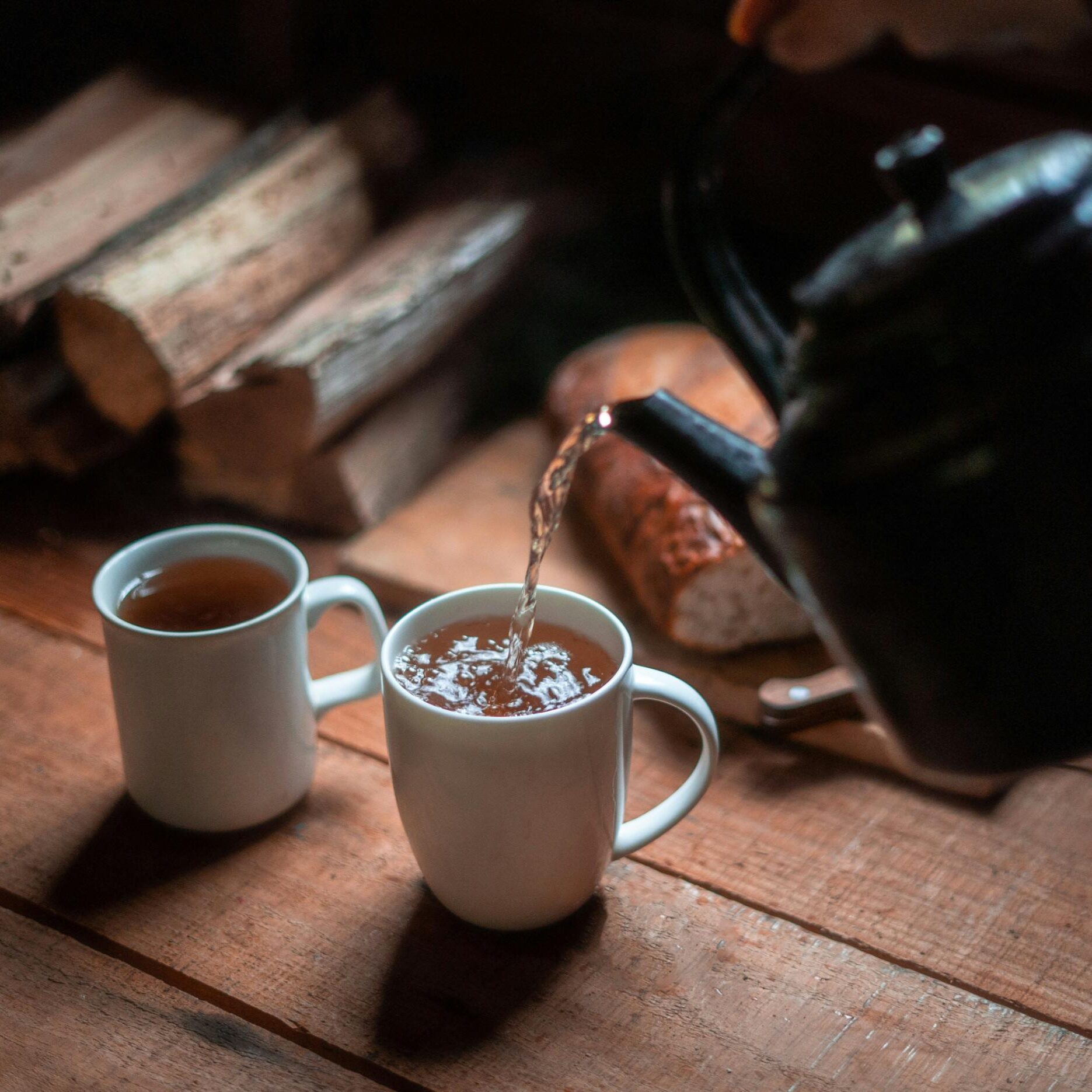 A black kettle pours a warm, amber-colored liquid into one of two white mugs resting on a rustic wooden table. The cozy, dimly lit scene is set against a background of stacked firewood and a loaf of bread.