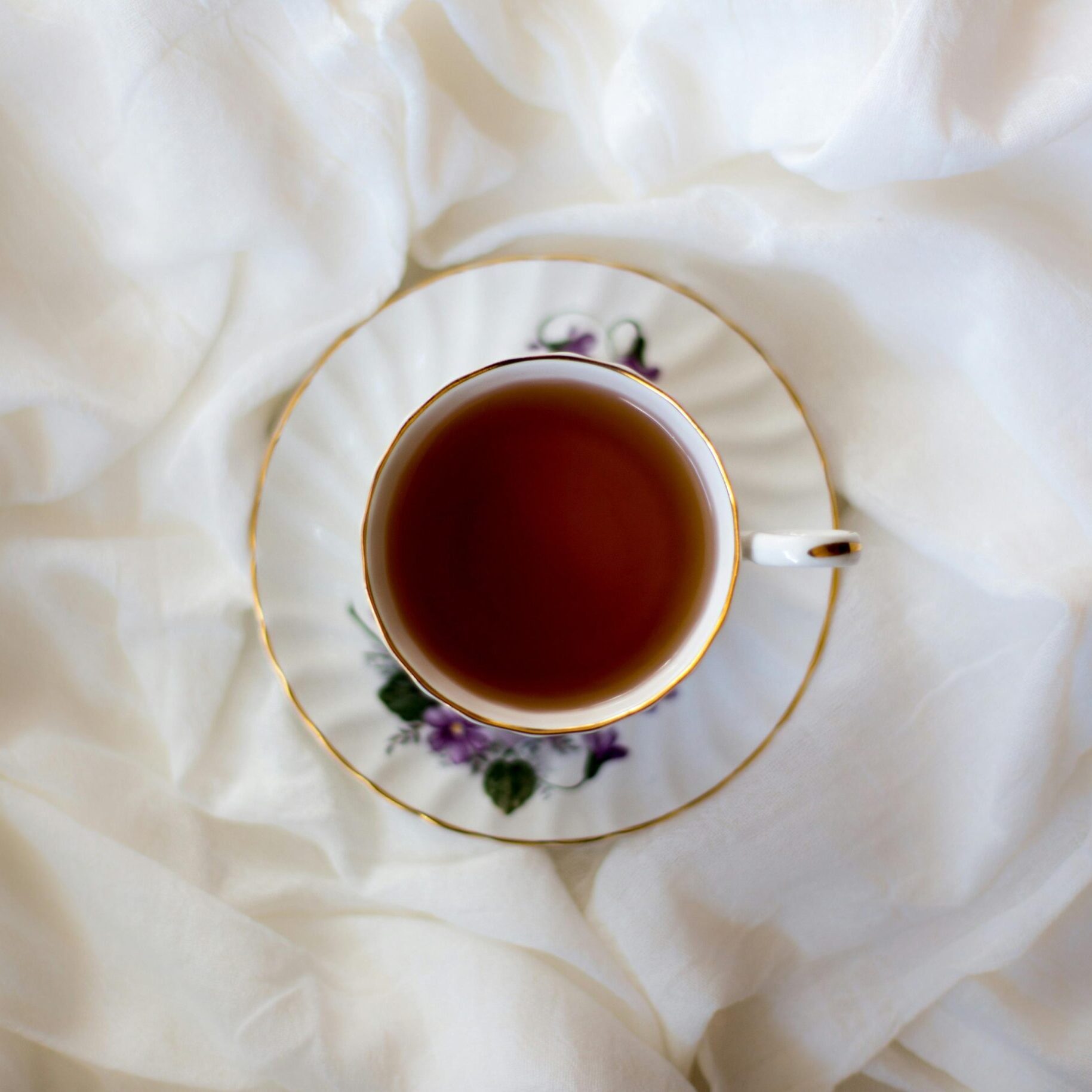 A delicate porcelain teacup filled with dark tea sits centered on a matching floral saucer. The set is nestled within the soft, rippling folds of a crisp white fabric, creating a clean and serene aesthetic.