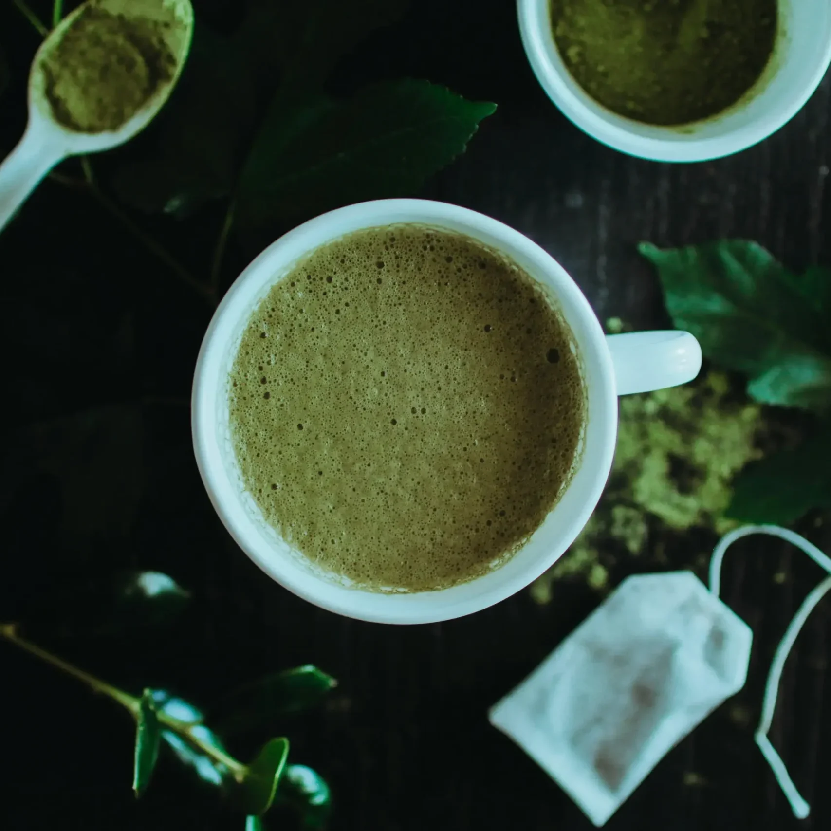 An overhead shot features a frothy cup of green matcha tea centered among loose powder, a tea bag, and fresh leaves. The dark wooden background and soft lighting create a moody, organic atmosphere for the tea preparation.